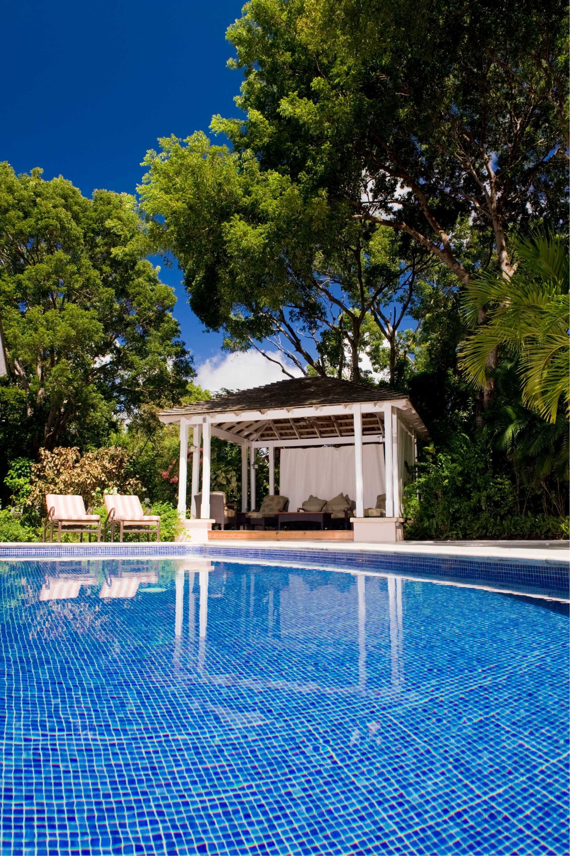 Swimming pool with blue tiled surface, white gazebo, lounge chairs, and lush green trees against a blue sky.