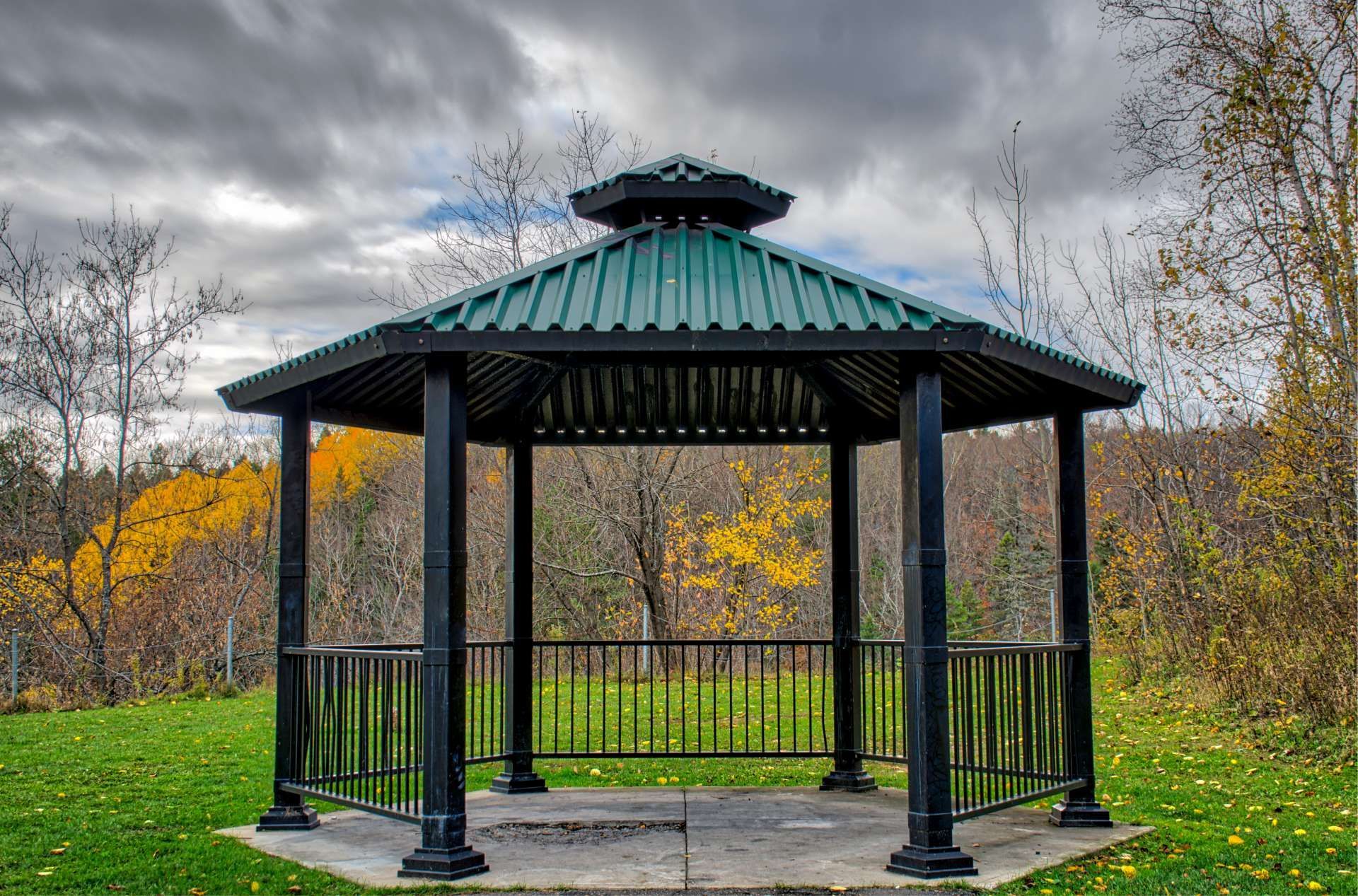 Black gazebo with green roof in a grassy park setting, autumn foliage in background, cloudy sky.