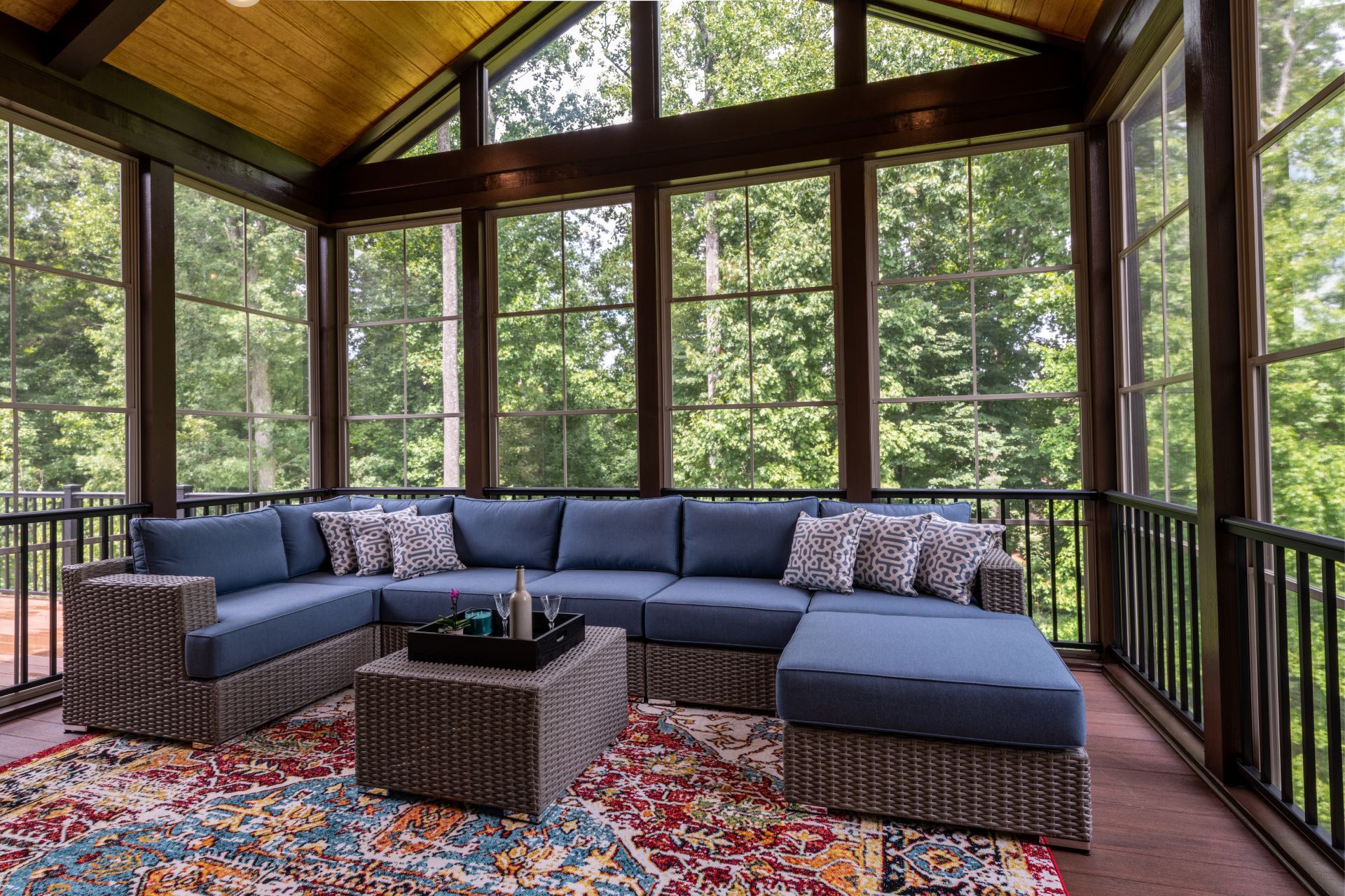 Screened porch with blue sectional, patterned rug, surrounded by windows with a forest view.