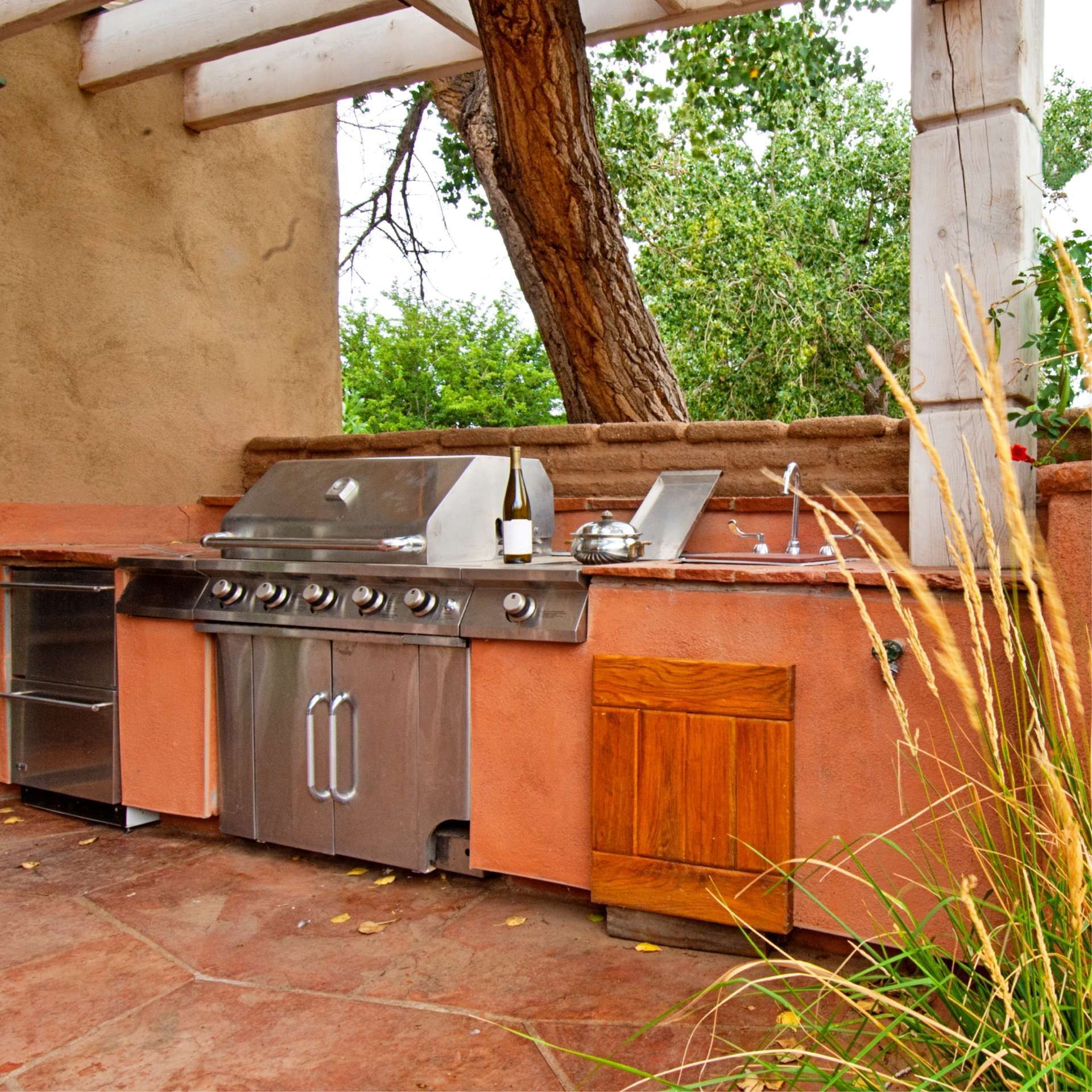 Outdoor kitchen with stainless steel grill, red stucco, tree trunk.
