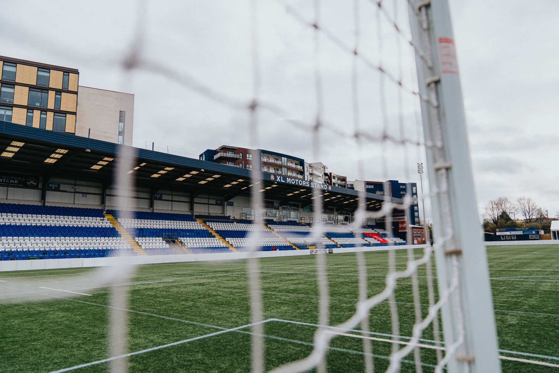 A football field with a goal net in the foreground and a stadium in the background.