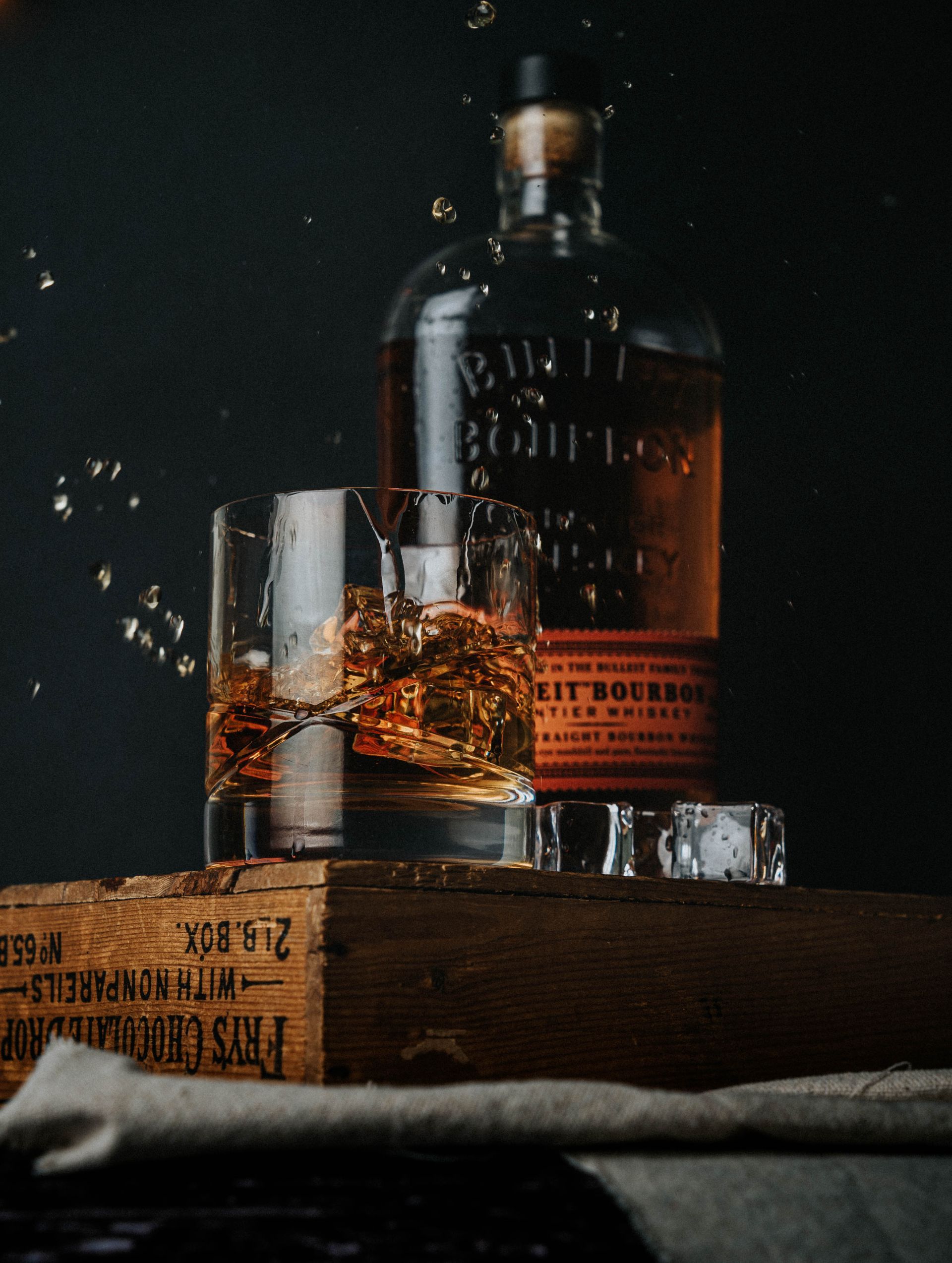 A bottle of whiskey next to a glass of whiskey with whiskey drops out of the glass 