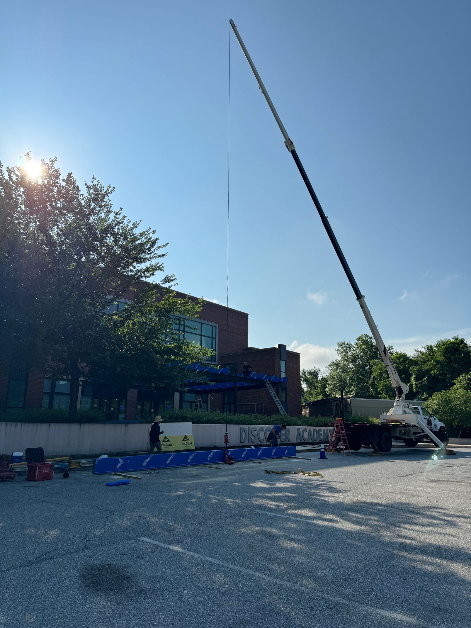 Crane lifting a tall antenna near a brick building on a sunny day.