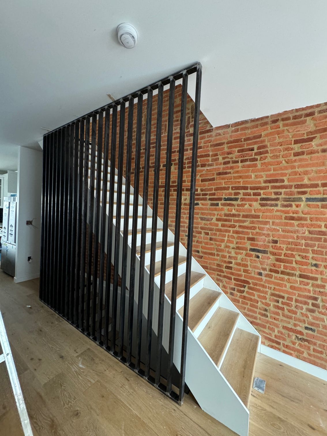Black vertical metal slat wall next to white staircase with light wooden steps, brick wall background.
