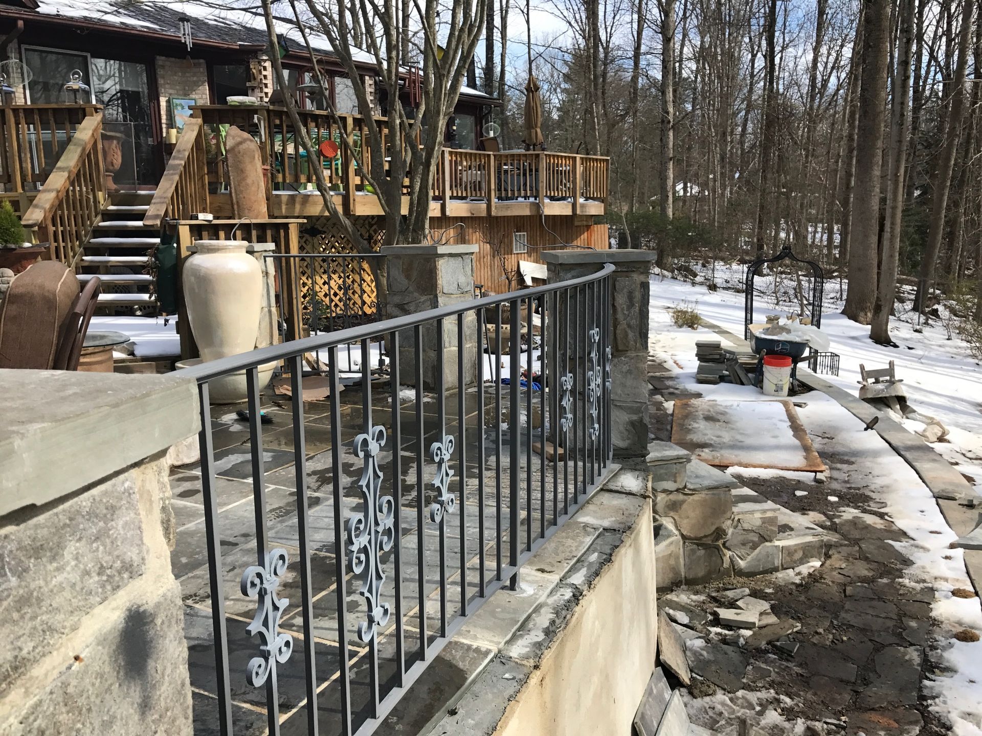 Stone patio with metal railing, steps up to a wood deck, snow-covered ground, and a wooded backdrop.