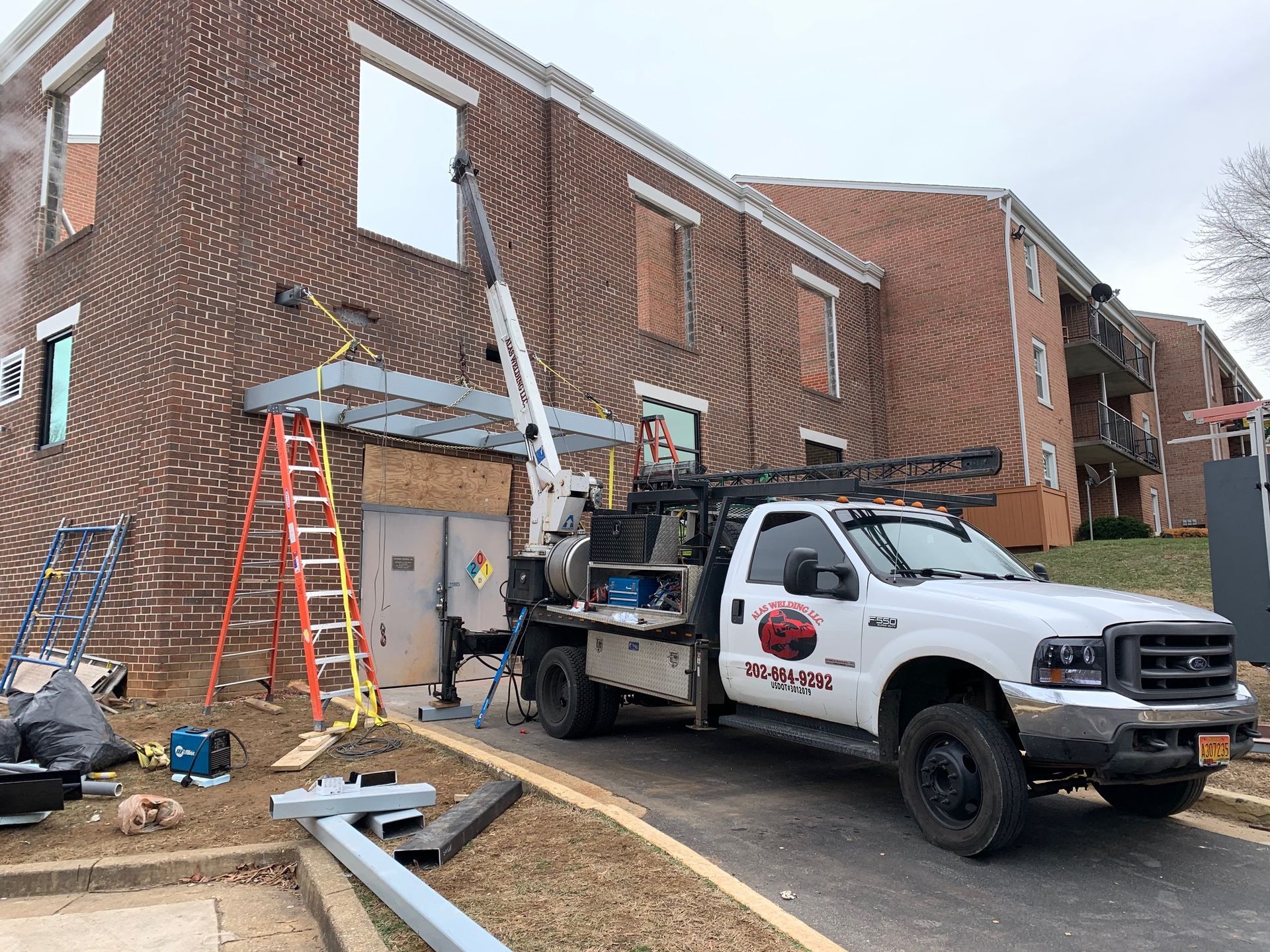 White truck with crane next to brick building, construction site. Ladder, boards, and metal parts present. Overcast sky.