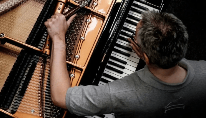A piano technician tuning a grand piano, using a tuning hammer to adjust the strings while pressing keys to test the pitch