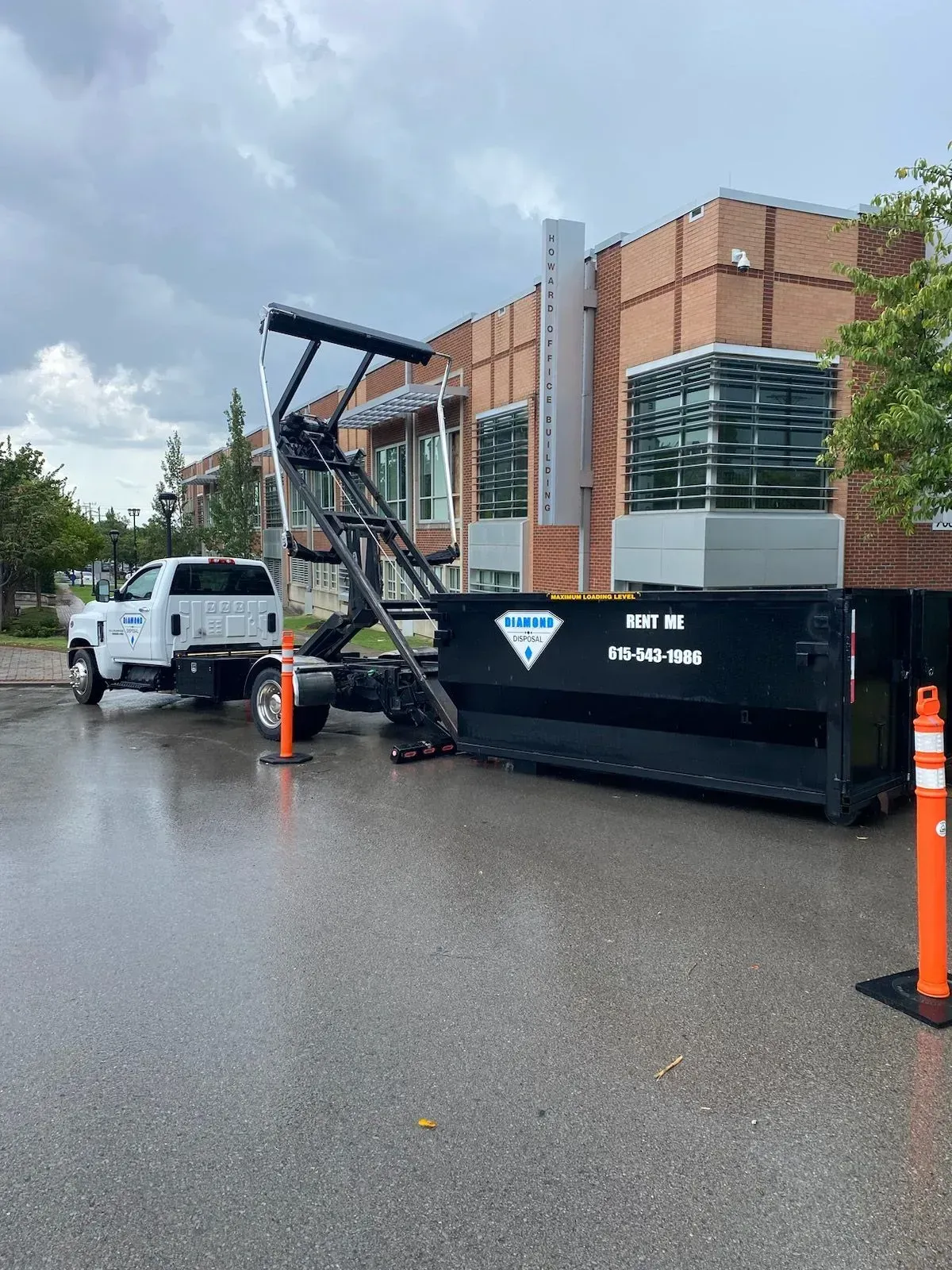 A white truck with a mechanical arm is parked in a parking lot, loading or unloading a large black waste dumpster.