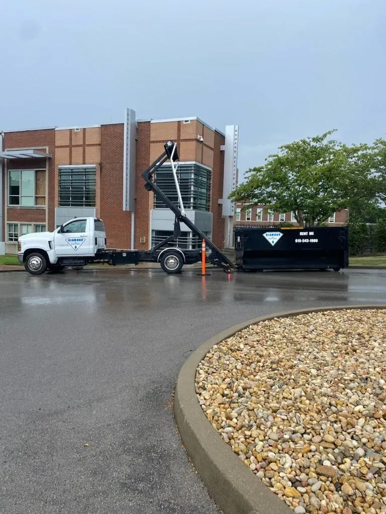 A white truck with a hydraulic arm lifting a large black dumpster in a paved parking lot in front of a brick building.