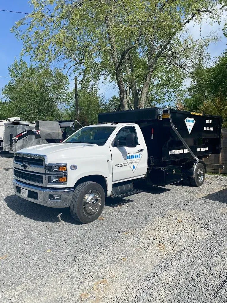 A white Chevrolet dump truck with a black trailer parked on a gravel lot under a sunny sky.