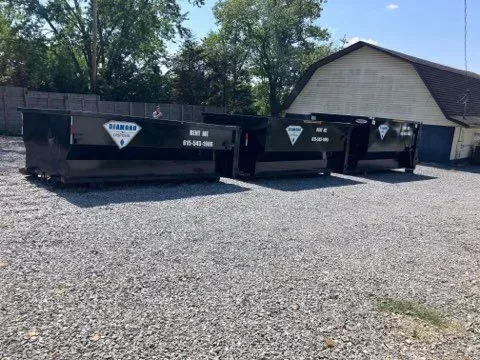 Three black dumpster bins with white diamond-shaped logos lined up on a gravel lot next to a barn-style building.
