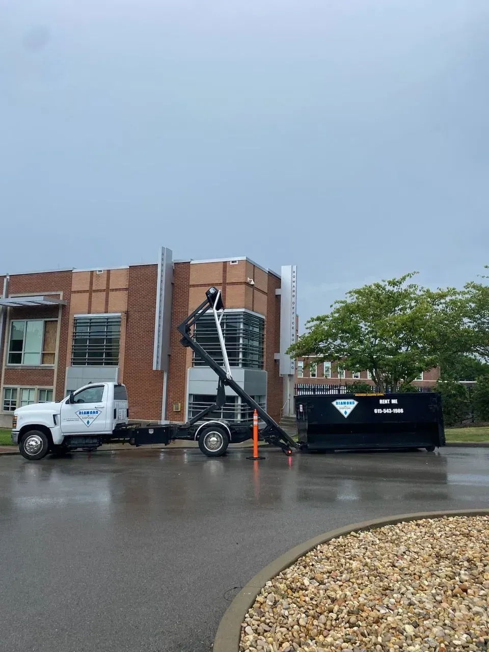 A white roll-off truck is parked in a wet parking lot, dropping a black dumpster in front of a brick building.