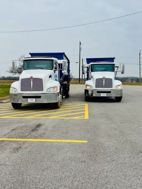 Two white Kenworth semi-trucks with blue dump trailers parked side-by-side on an asphalt lot.
