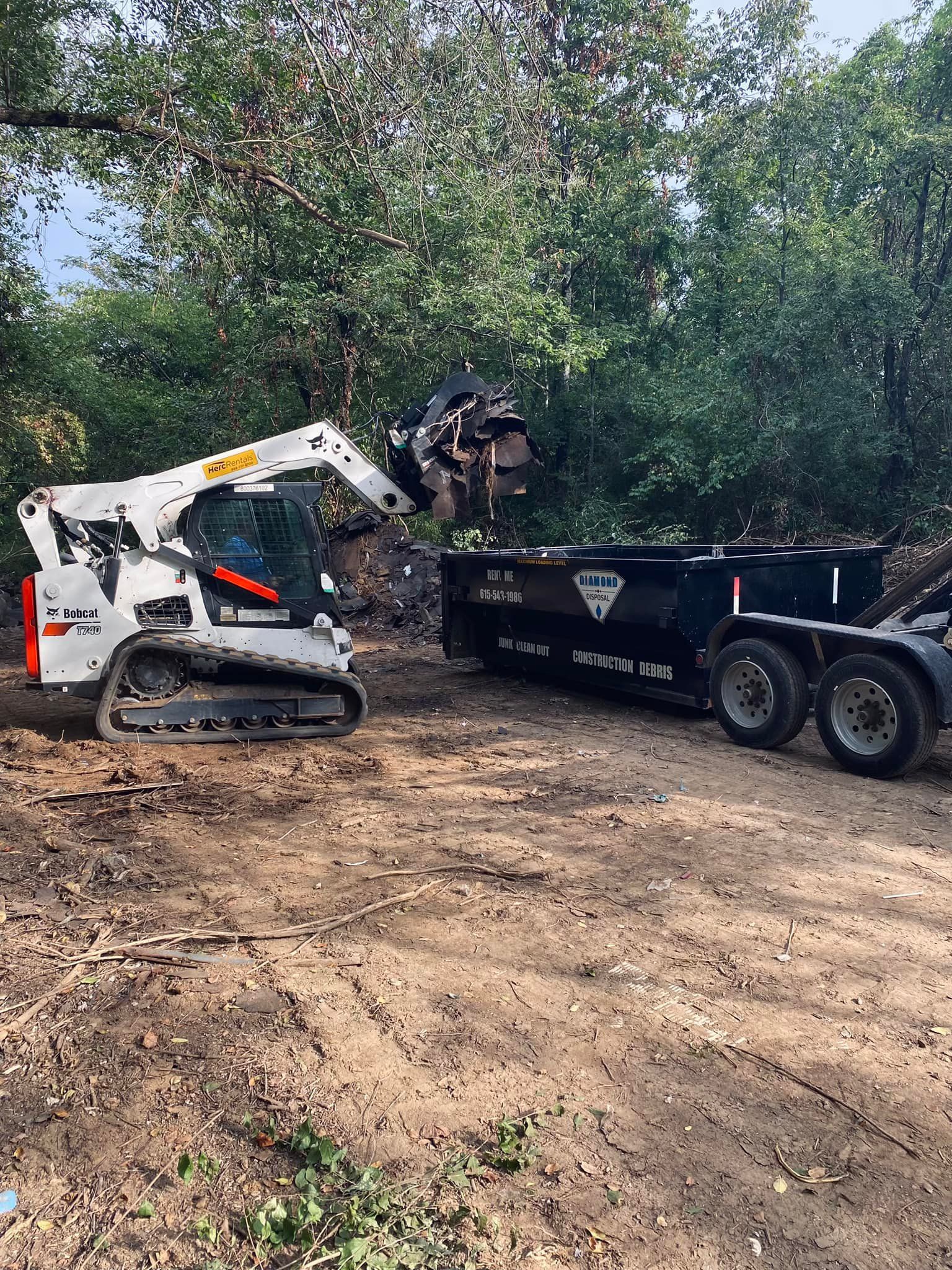 A white skid-steer loader uses a grapple attachment to load brush into a black dumpster trailer in a wooded area.