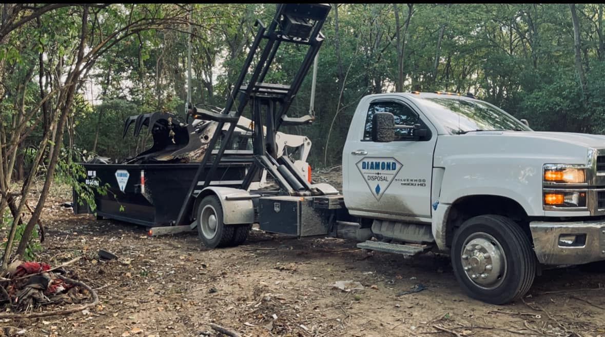 A white truck with a large, black dumpster container hooked to its rear, parked in a wooded area.