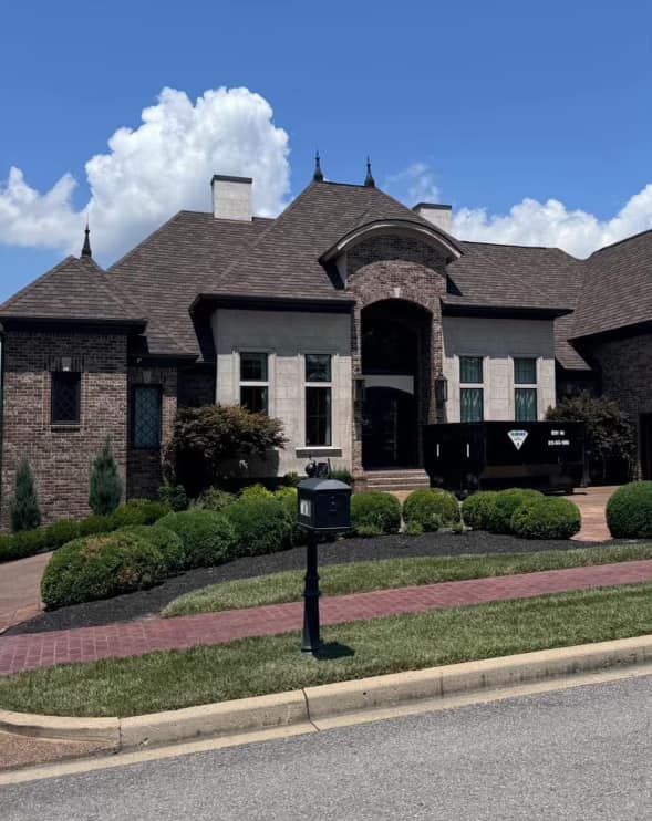 A large brick and stone house with a dark roof and landscaping in the front yard under a blue sky.