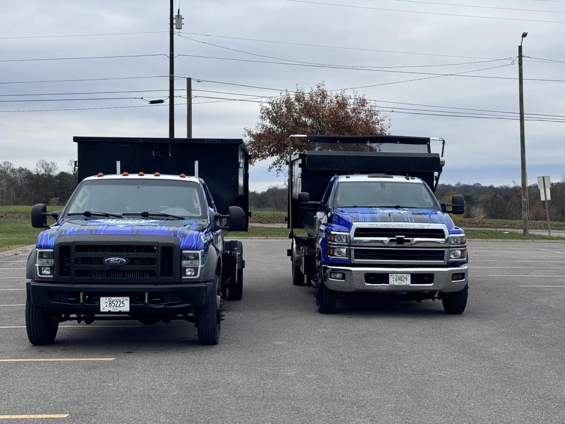 Two blue dump trucks parked side-by-side in a large, paved parking lot under a cloudy sky.