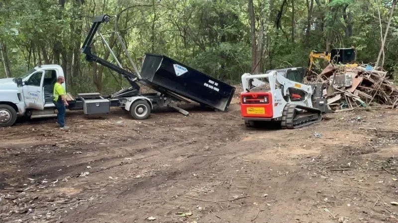 A person stands by a roll-off dump truck and a Bobcat loader clearing debris in a wooded area.