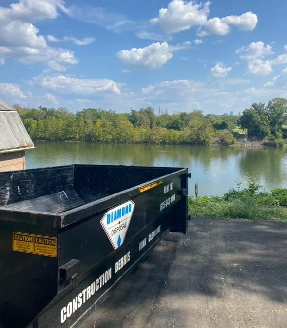 A large black Diamond Disposal dumpster sits on a paace overlooking a calm river surrounded by trees under a blue sky.