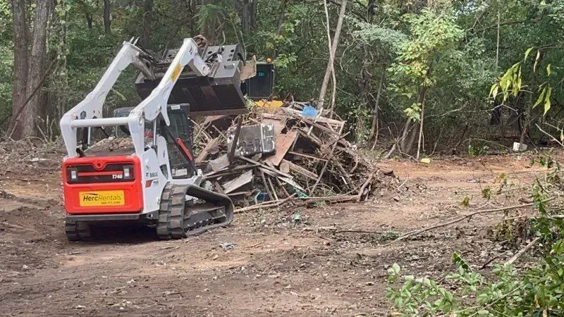 A white skid steer loader uses a grapple attachment to clear a large pile of scrap metal and debris in a wooded area.