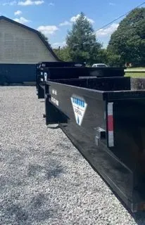 A long, black roll-off dumpster sitting on a gravel lot next to a building.