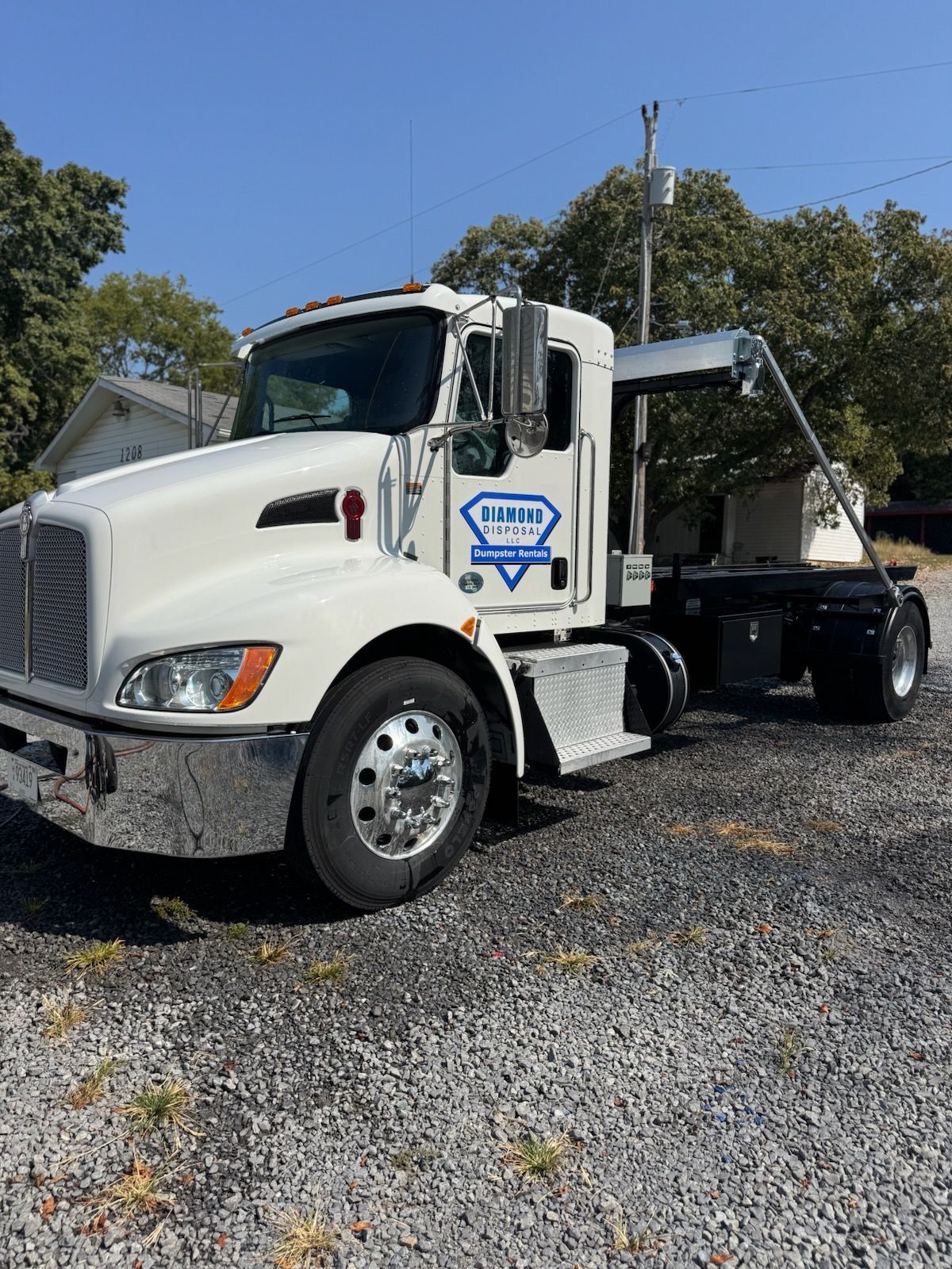 A white roll-off truck with a blue diamond logo on the door, parked on a gravel lot under a clear blue sky.