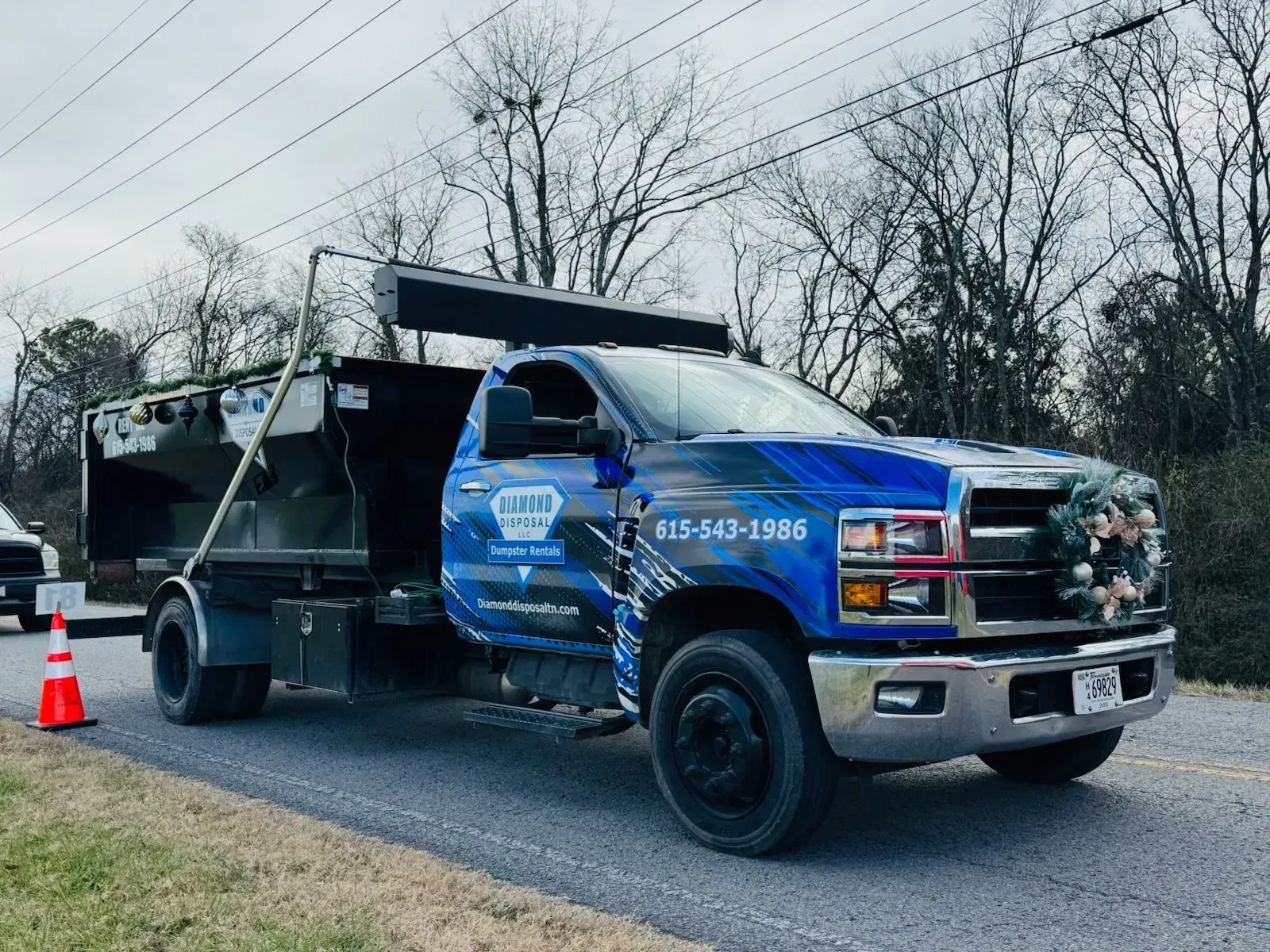 Blue service truck with a dump bed and Christmas wreath on the grille parked on a gravel road with a traffic cone nearby.