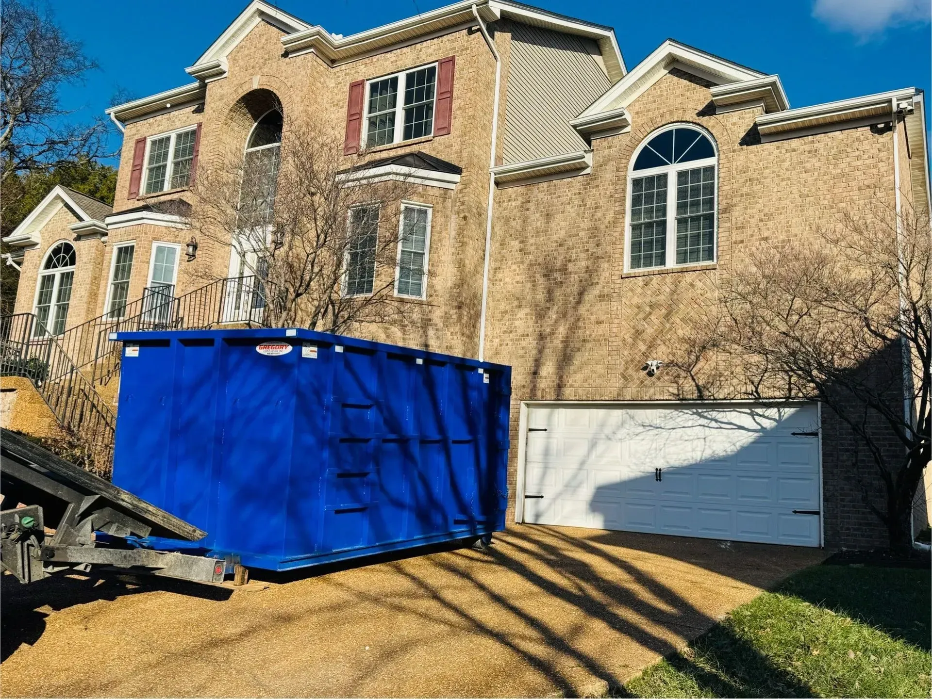 A large, bright blue dumpster sits on a gravel driveway in front of a tan brick house with a two-car garage.