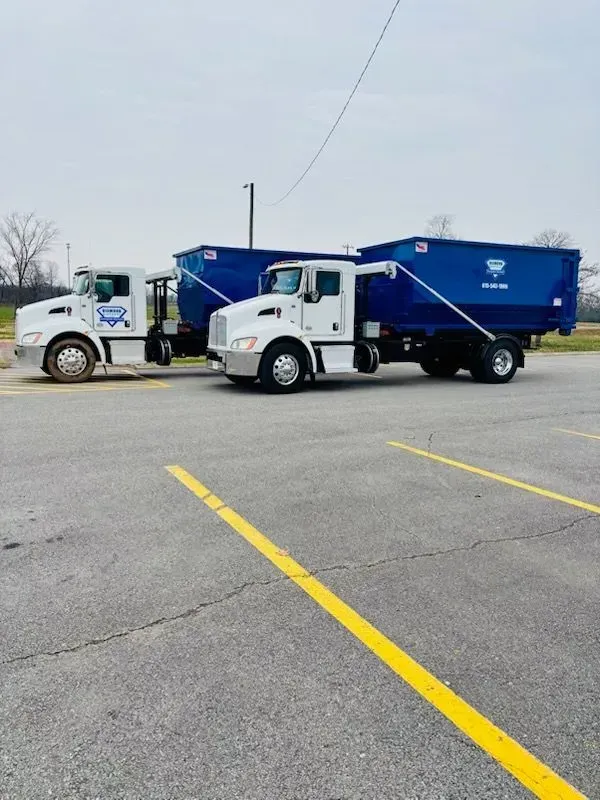 Two white roll-off trucks with blue dumpsters parked side-by-side in a paved lot.