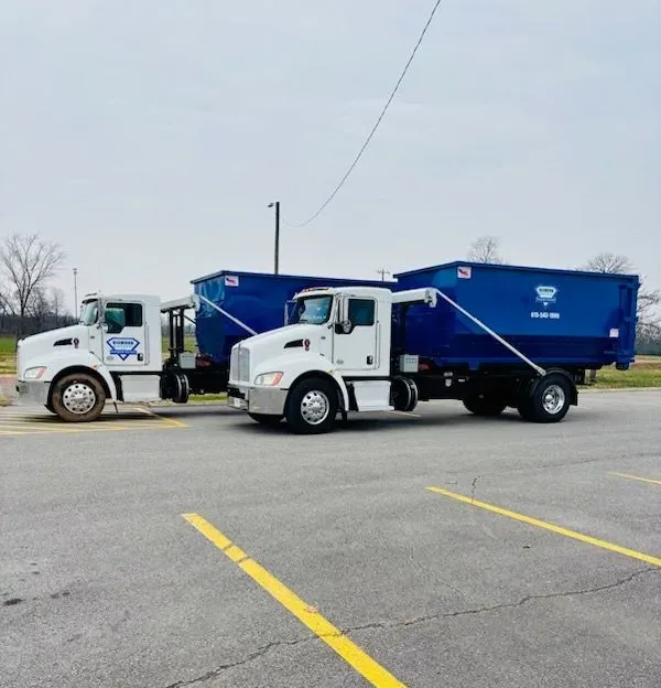 Two commercial dump trucks, one blue and one black, parked side-by-side on an asphalt lot under an overcast sky.