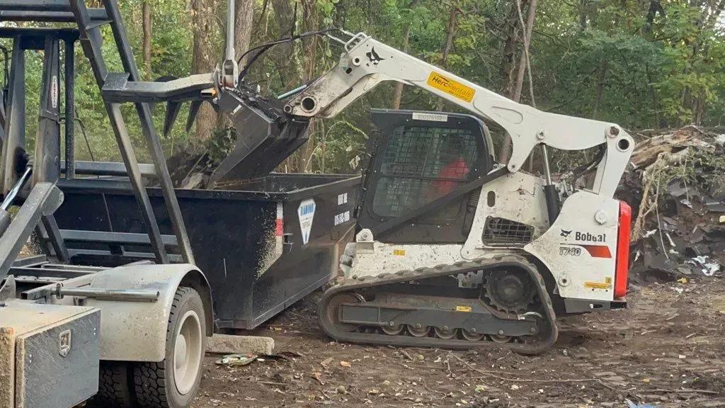 A white Bobcat compact track loader loads debris into the back of a black dump trailer in a wooded area.