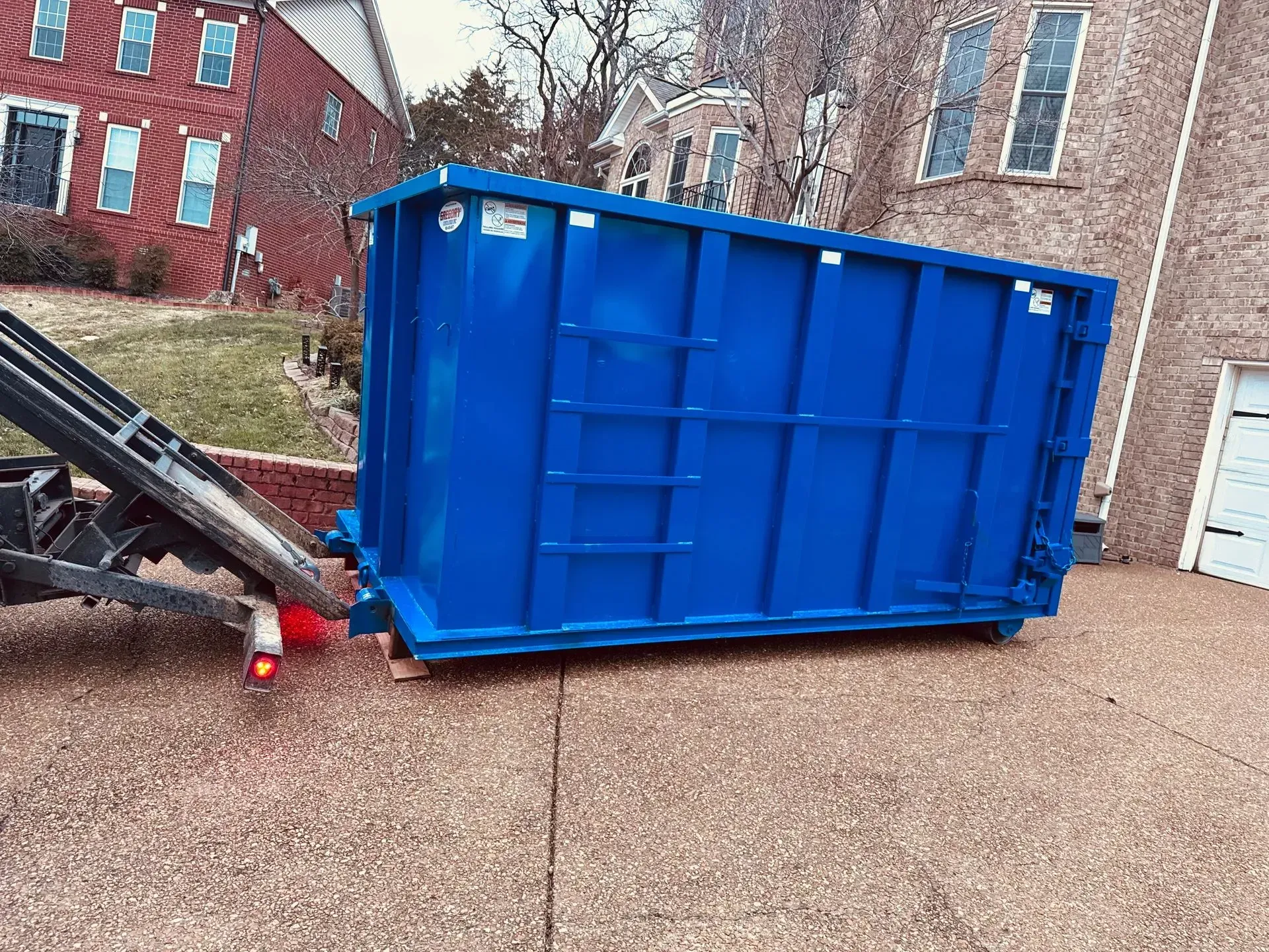 A bright blue roll-off dumpster being unloaded from a trailer onto a residential stone driveway.