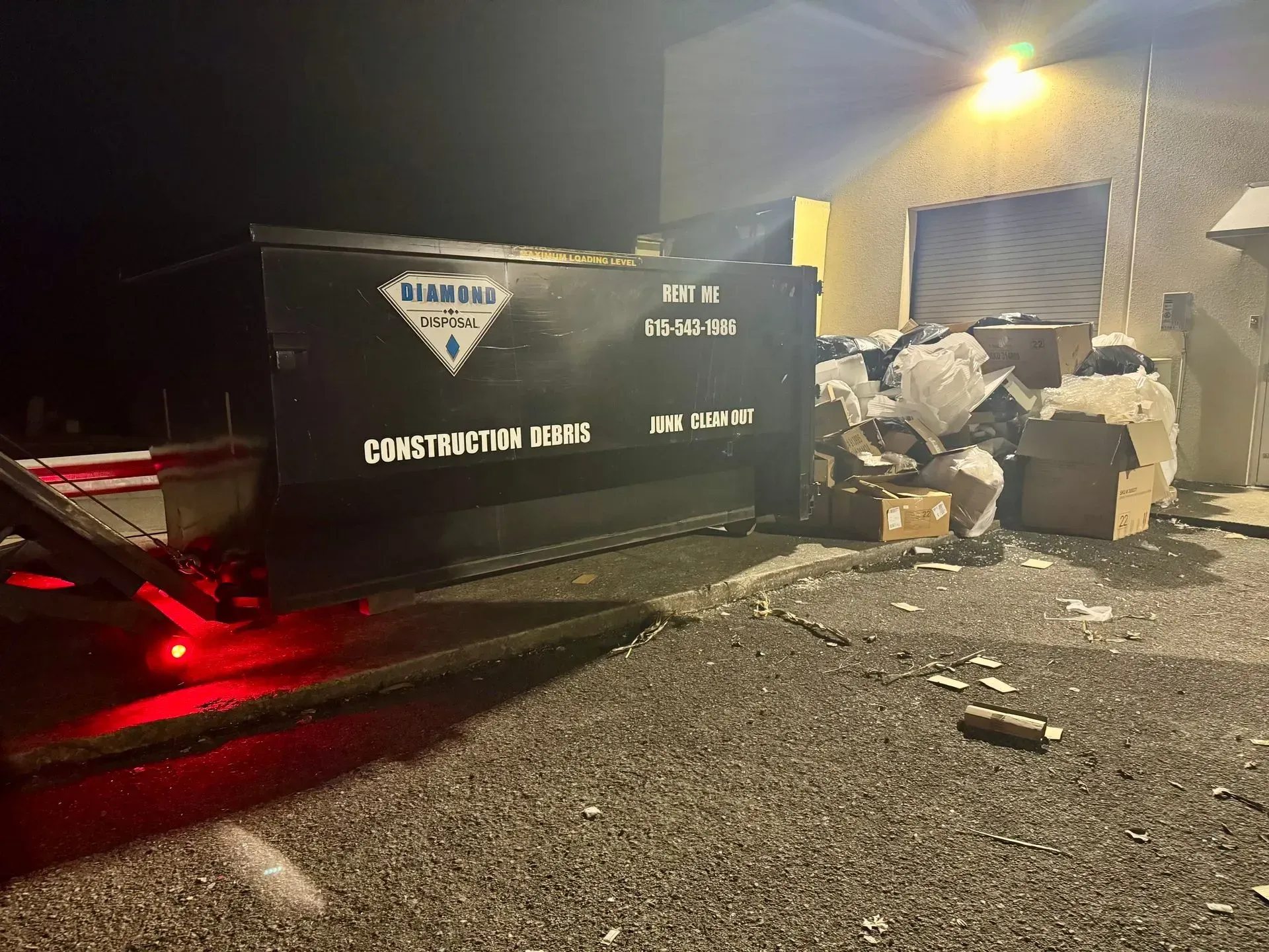 A black construction dumpster sits in a parking lot at night, surrounded by overflowing trash and cardboard boxes.
