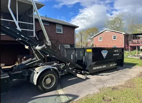 A black roll-off dumpster being lowered onto a driveway by a truck next to a brick house under a blue sky.