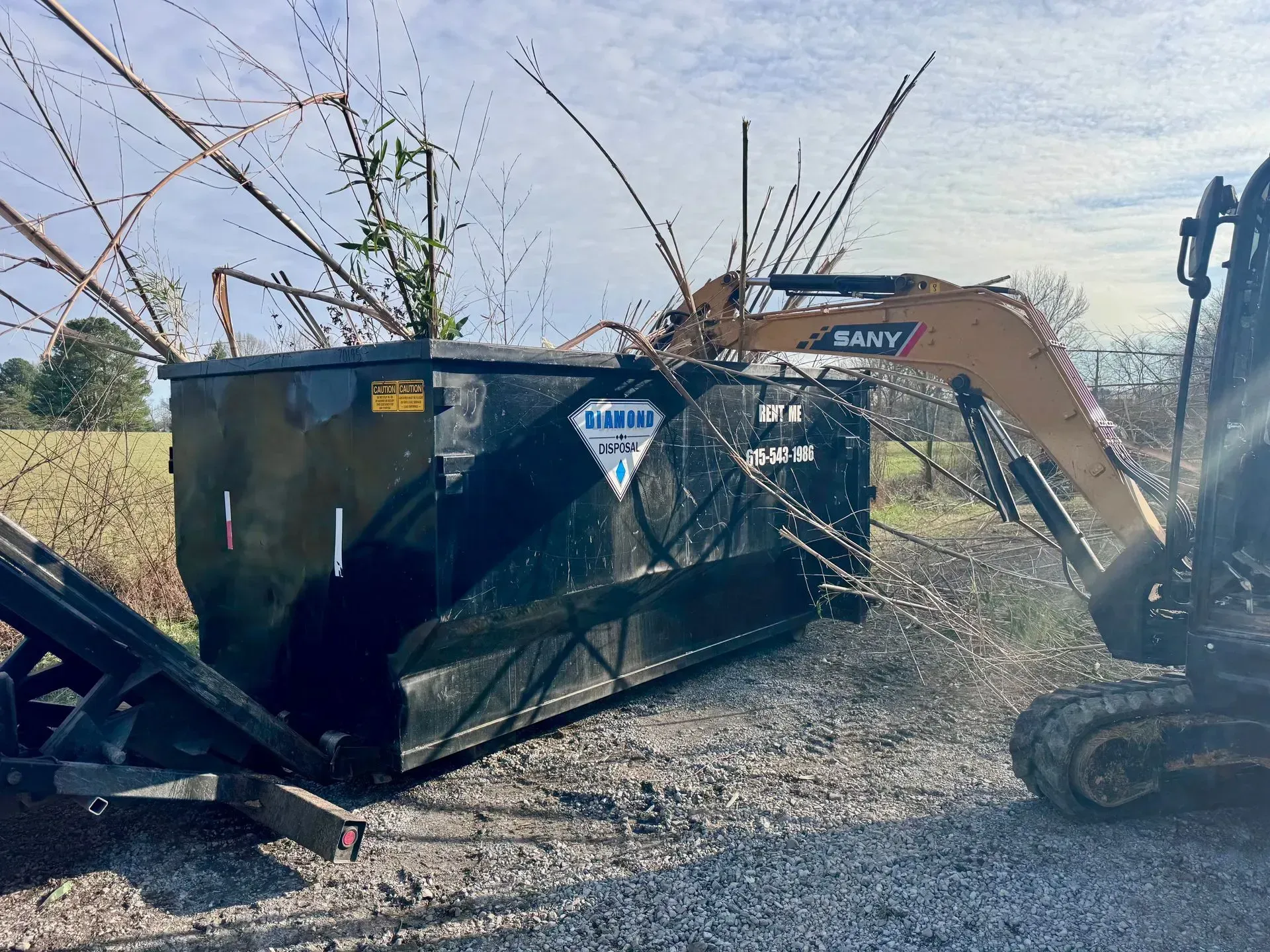 An excavator arm loading tree branches into a large, black dumpster on a gravel surface.