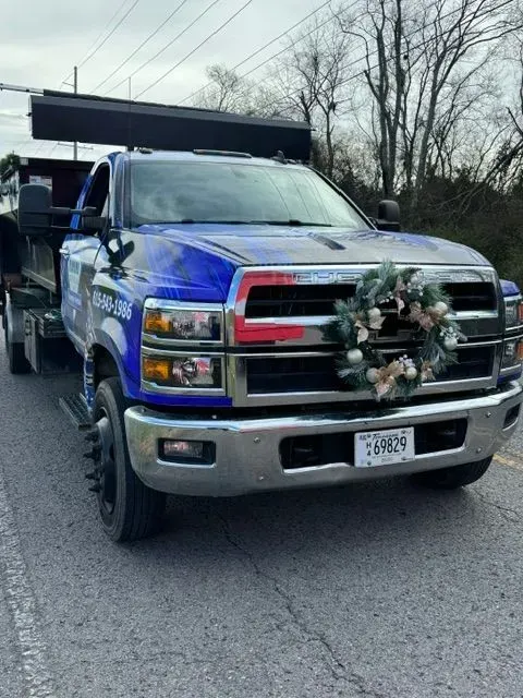 A blue commercial dump truck on a road with a festive holiday wreath attached to the front grill.