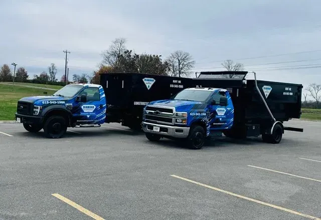 Two blue work trucks with black dump trailers parked side-by-side in a paved lot.