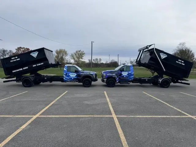 Two dark-colored dump trucks parked facing each other in an empty parking lot, both with their beds tilted upward.