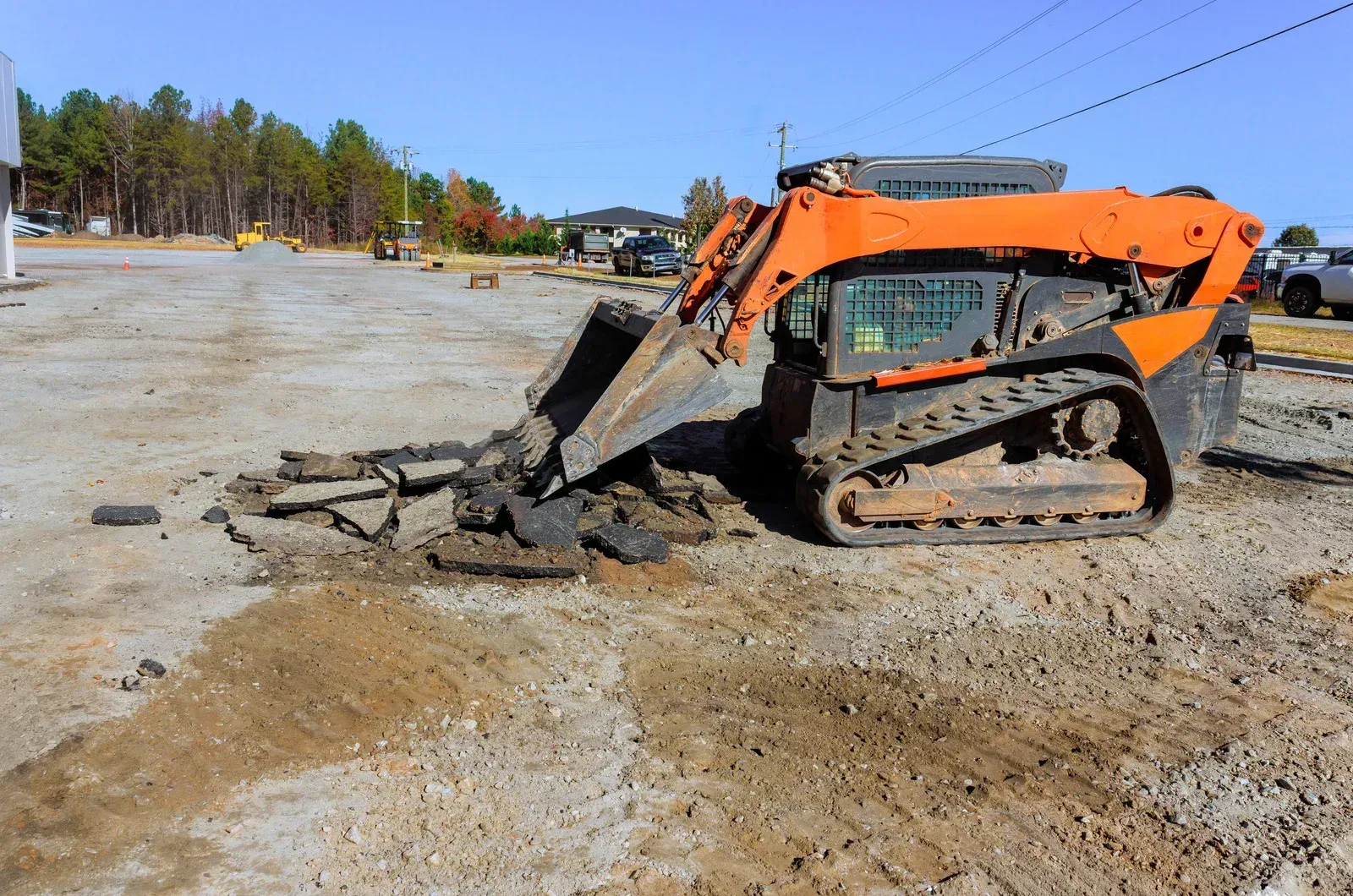 An orange skid steer loader scoops up broken asphalt on a gravel lot under a clear blue sky.