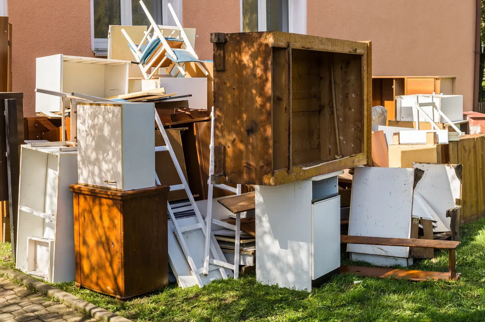 A pile of discarded wooden and white furniture, including cabinets, chairs, and shelves, sitting on a patch of grass.