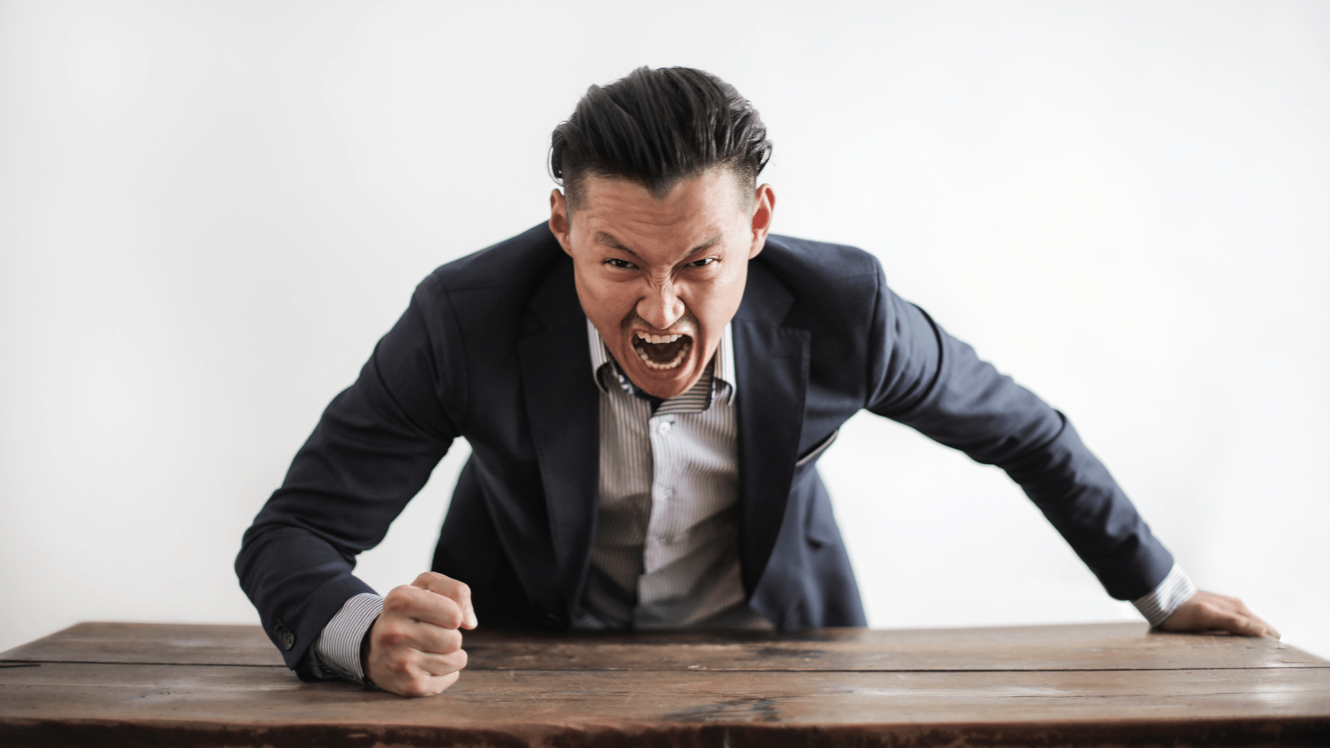Angry businessman in suit leaning over table, shouting at camera with clenched fists and expressive face.