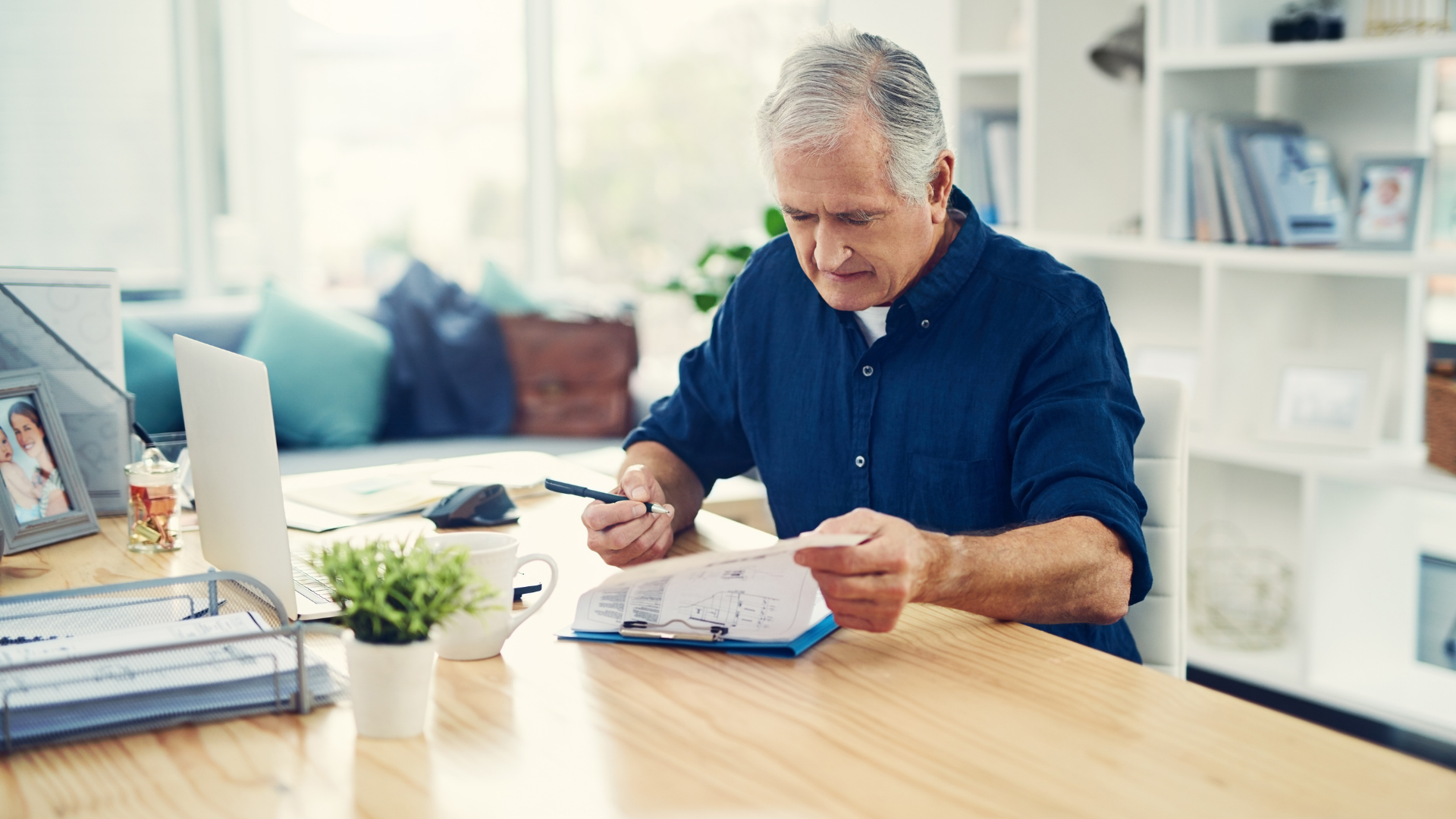 Landlord in blue shirt writes in notebook at desk with laptop and photos, in a bright, plant-decorated office.