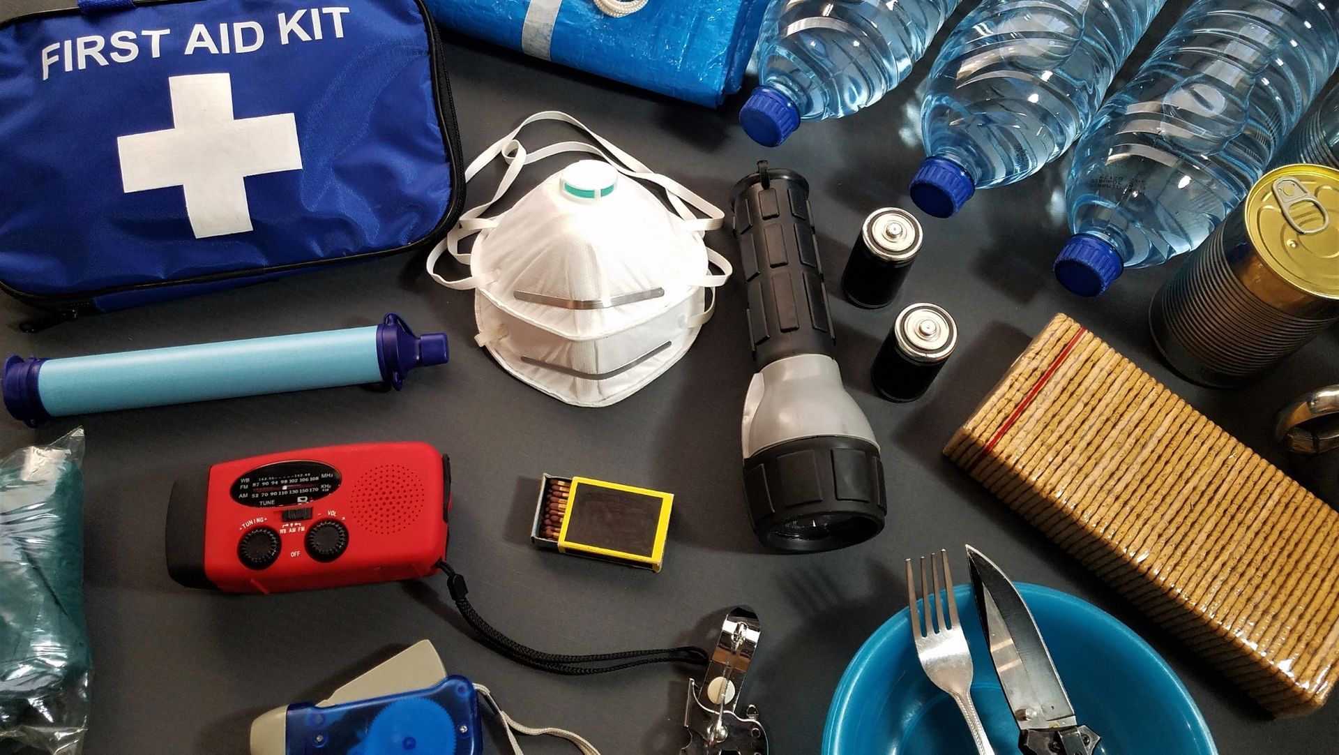 Emergency supplies on table including first aid kit, water, masks, flashlight, canned food, radio, and utensils.