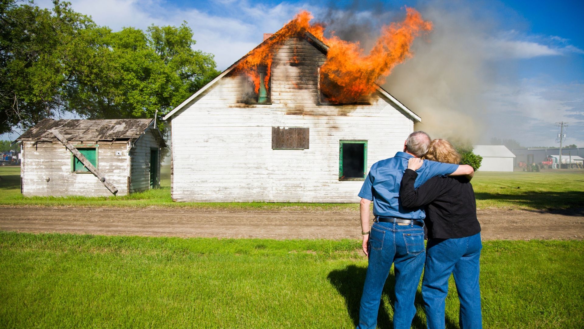 Older couple stands arm in arm watching white house with flames and smoke from upstairs window, surrounded by greenery.