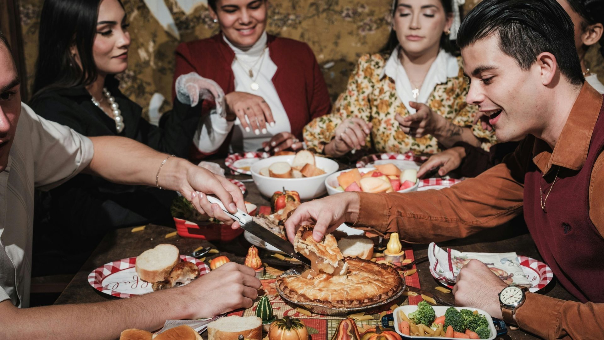 A group of people sit around a table sharing food, including a pie, bread, fruit, and vegetables. They are smiling, reaching for food, and appear to be enjoying a festive meal together.