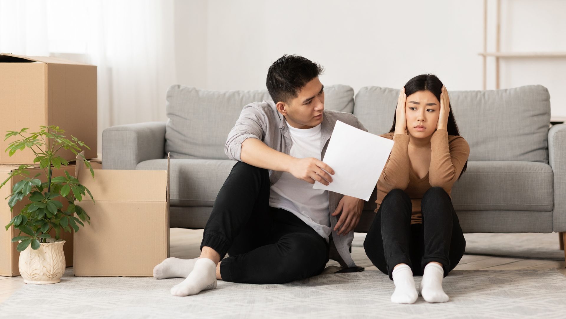 Man and woman sitting by unpacked boxes, looking concerned, indicating a move or relocation.