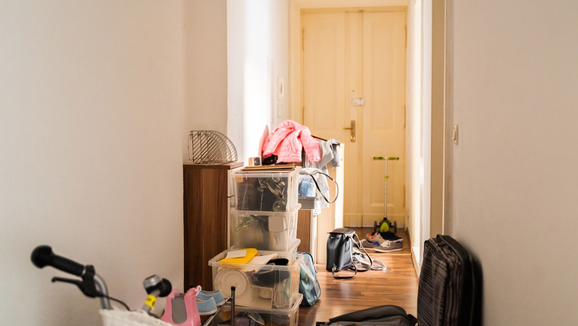 Cluttered hallway with storage bins, bicycle wheel, shoes, bags, pink jacket, and scooter on wooden floor.