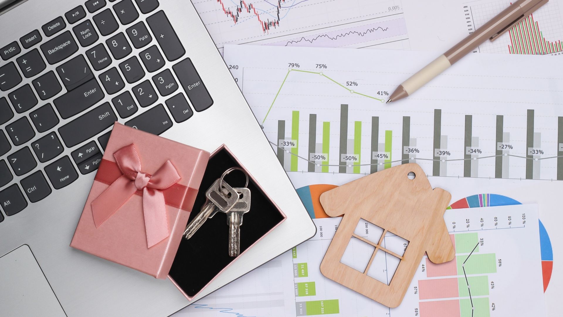 Laptop keyboard, pink gift box with house keys, wooden house keychain, and pen on financial charts and data sheets.
