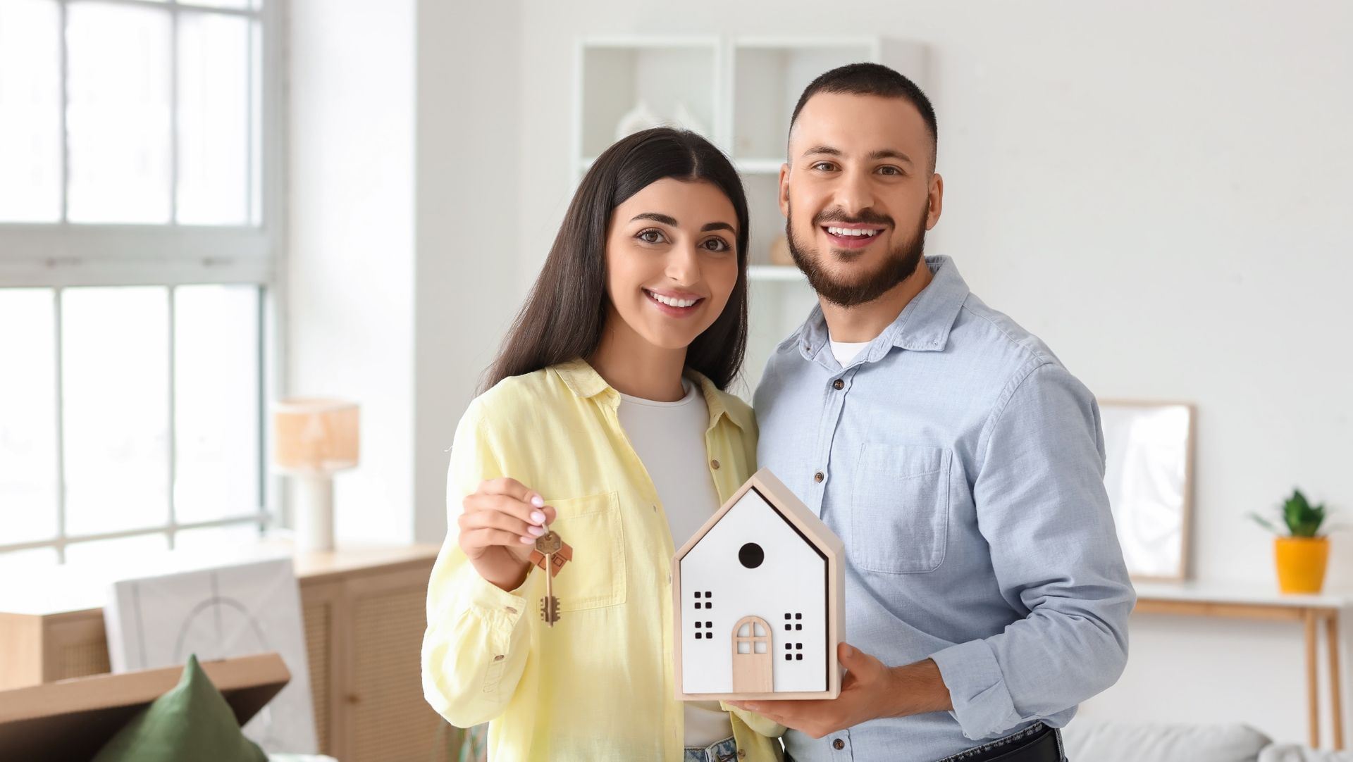 Smiling couple holding house model and keys in bright modern rental home, symbolizing new lease or move-in.