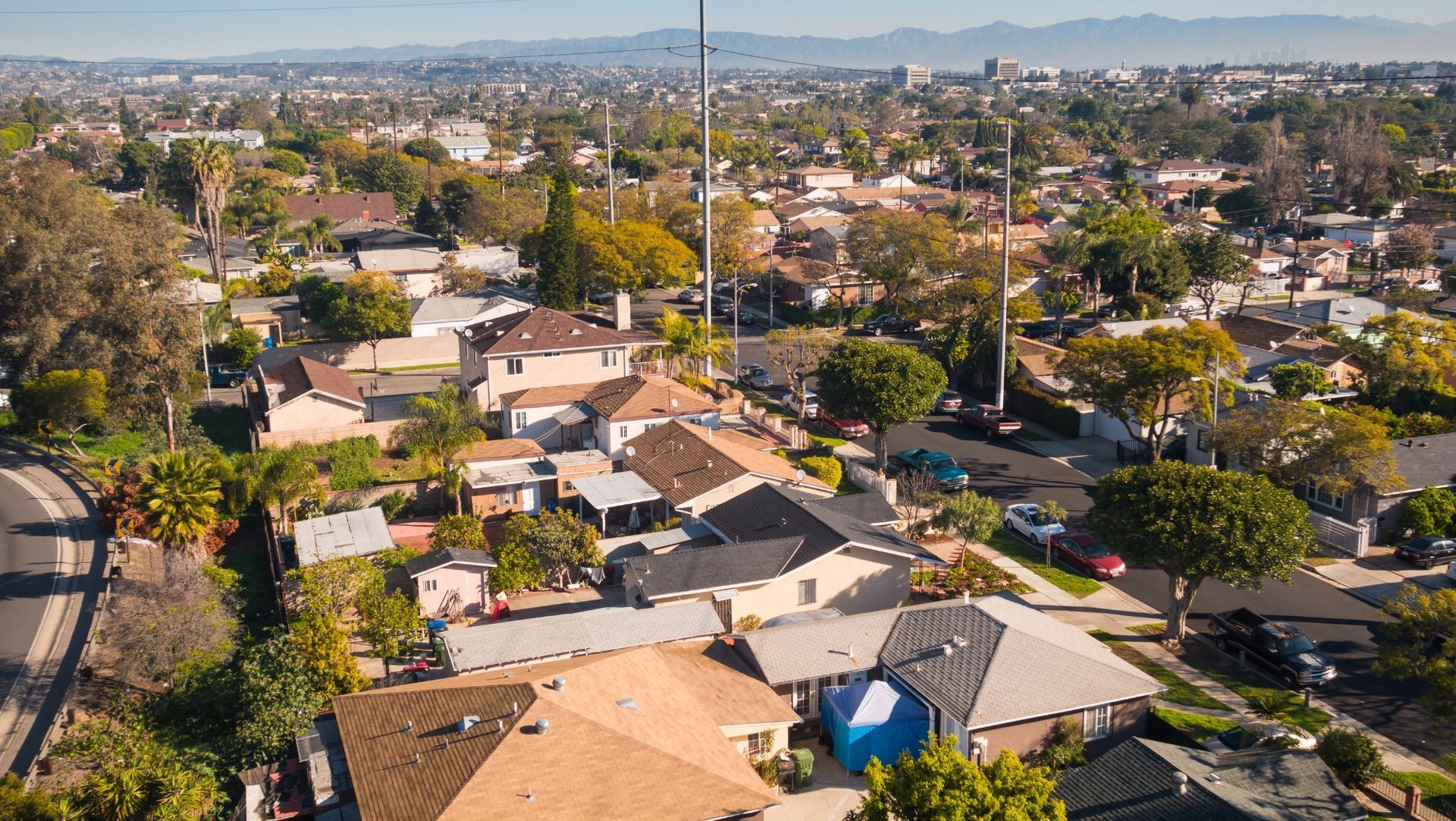 Aerial view of suburban neighborhood with single-family homes, tree-lined streets, distant city buildings, and mountains.